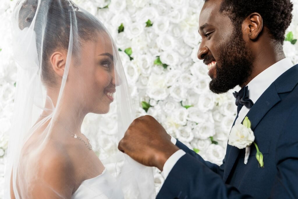happy african american bridegroom touching white veil and smiling near bride and flowers